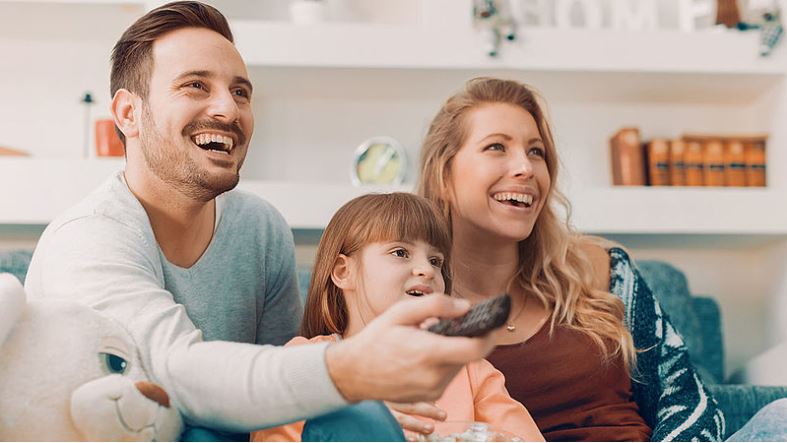 A couple and their kids are watching TV while the father is press buttons on a remote control